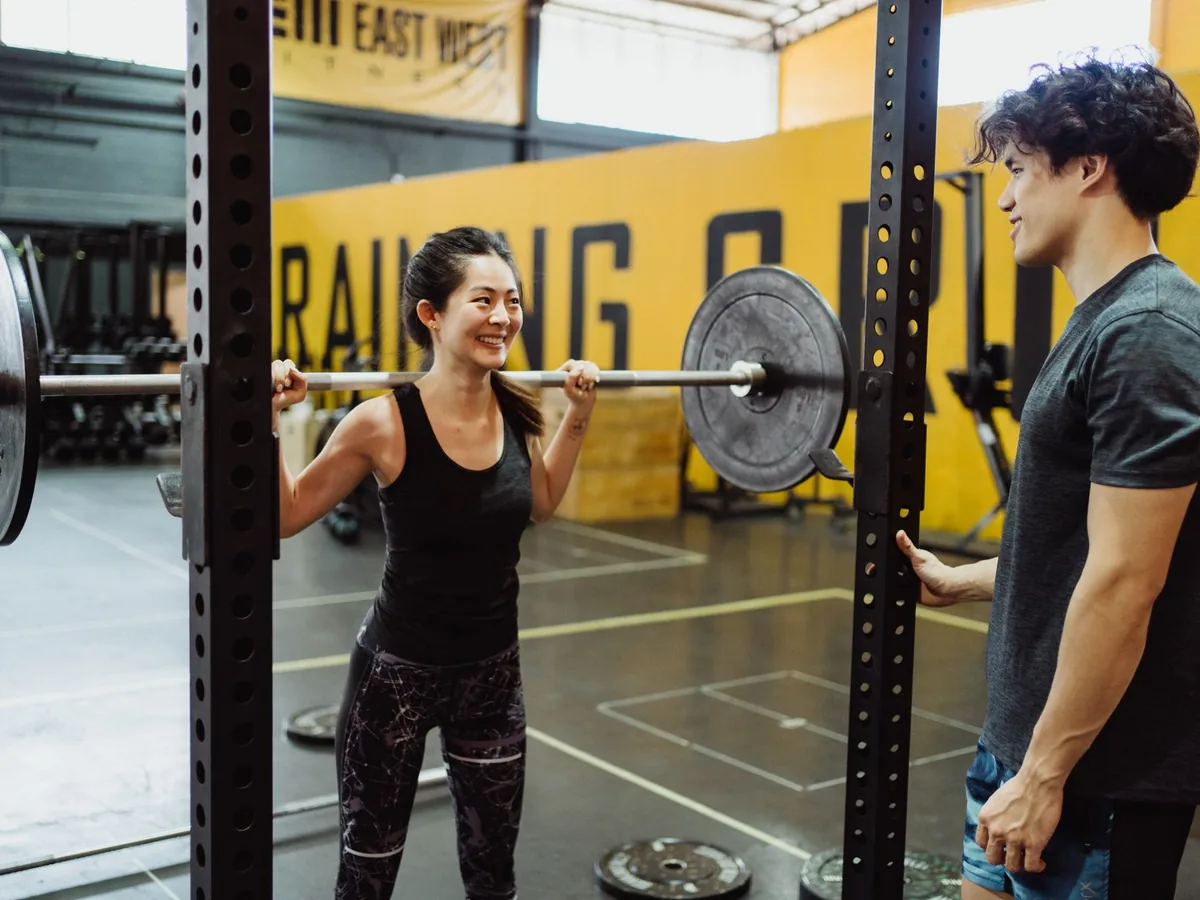 A woman lifts a barbell while a trainer watches beside her.