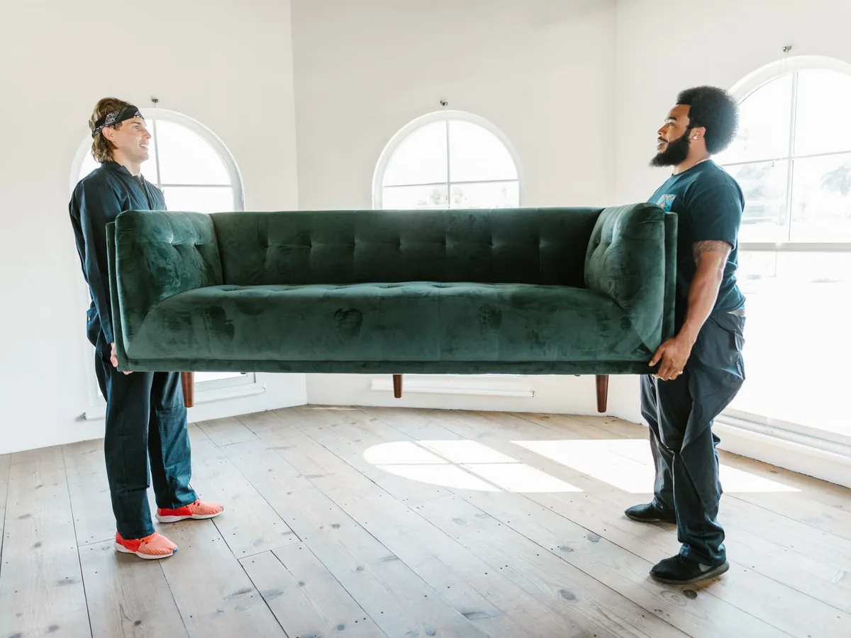Two removalists carry a green sofa in a bright room.