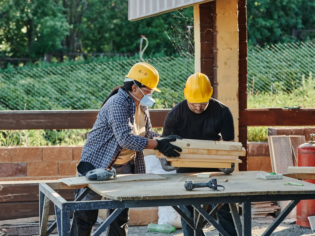 Two builders handle wooden planks beside a saw at an outdoor construction site.
